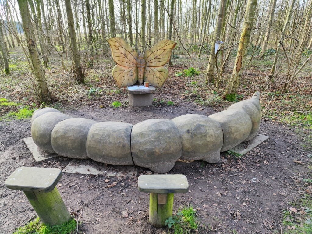 A large wooden sculpture of a caterpillar with butterfly wings, surrounded by a wooded area, featuring benches nearby.