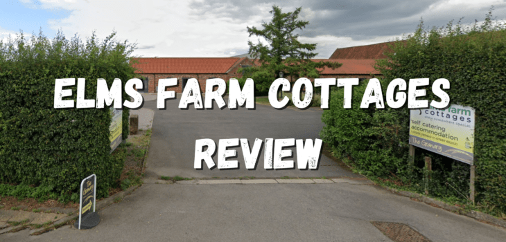 Entrance to Elms Farm Cottages features large white text reading "Elms Farm Cottages Review," bordered by hedges, with cottage signs and charming red-roofed buildings visible in the background beneath a cloudy sky.