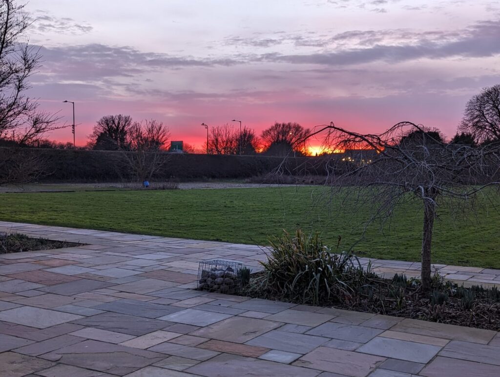 Sunset view over grassy field with stone patio in the foreground, featuring trees and street lights silhouetted against a colorful sky.