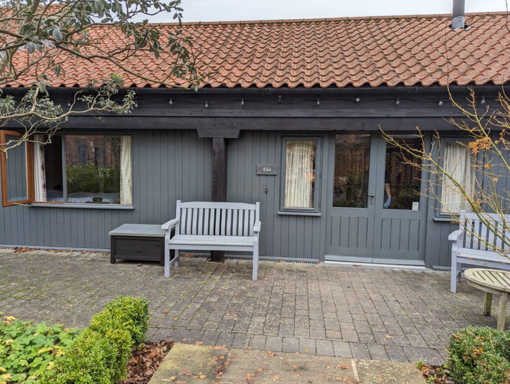 Exterior view of Elm cottage at Elms Farm Cottages, featuring grey wooden cladding, a tiled roof, and a small patio with seating.