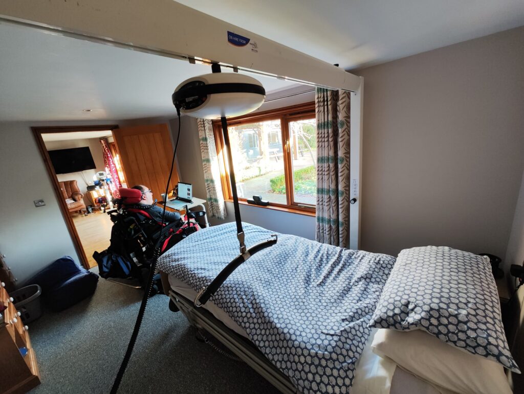 A bedroom in Elm Cottage featuring a bed with patterned bedding, a gantry hoist above it, and a view of the living area and garden through a window.