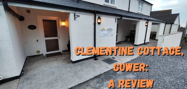 A white cottage with a covered entrance, outdoor lights, and a sign reading Clementine Cottage. Bold orange text on the image says, Clementine Cottage, Gower: A Review. Discover the charm of Gower’s beloved Clementine Cottage.