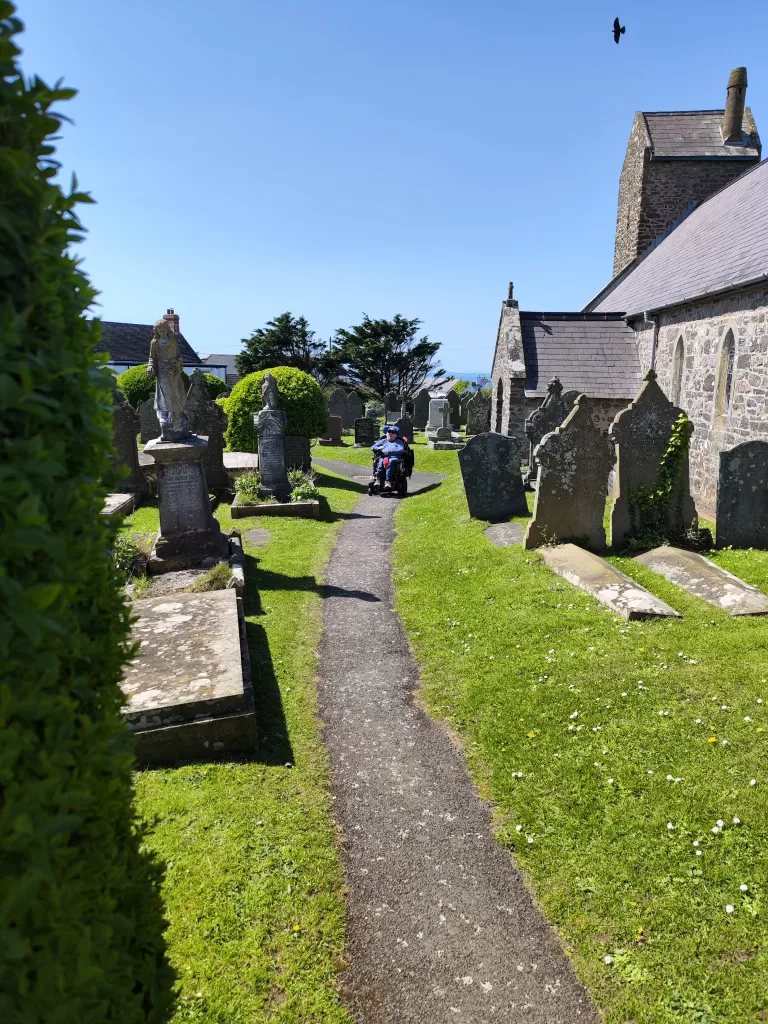 Person in a power chair navigating a grassy path in a cemetery with gravestones and a church in the background on a sunny day.