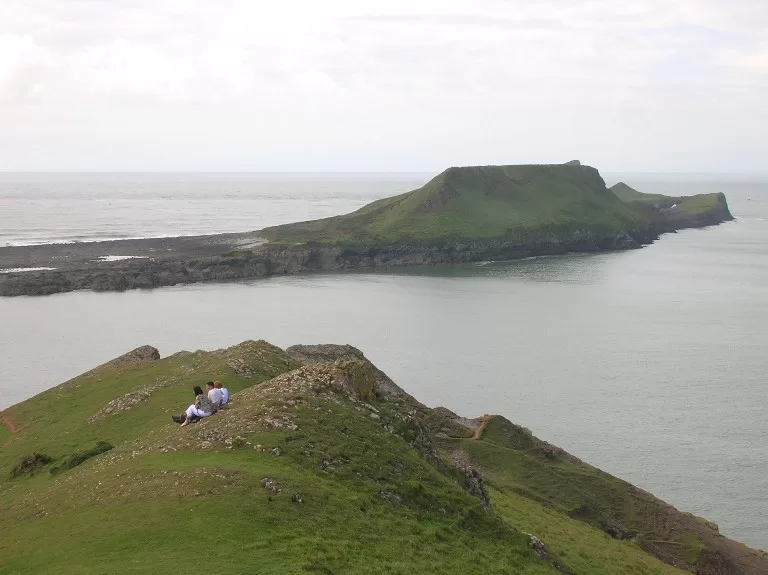 A scenic view of Worms Head from a nearby cliff, showcasing the green landscape and the coastline in Wales, with two people sitting on the grass.