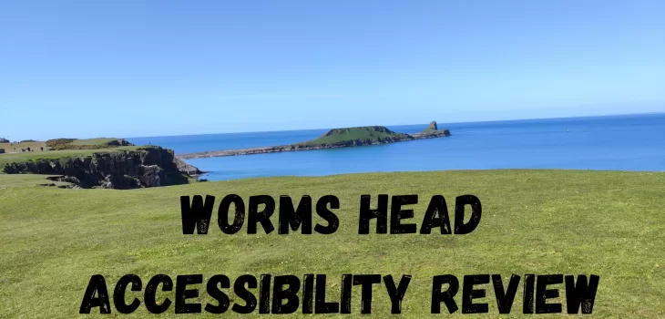 View of Worm’s Head, a grassy headland jutting into the blue sea under a clear sky, with bold black text reading “Worms Head Accessibility Review” over the green foreground, highlighting Worms Head accessibility for all visitors.