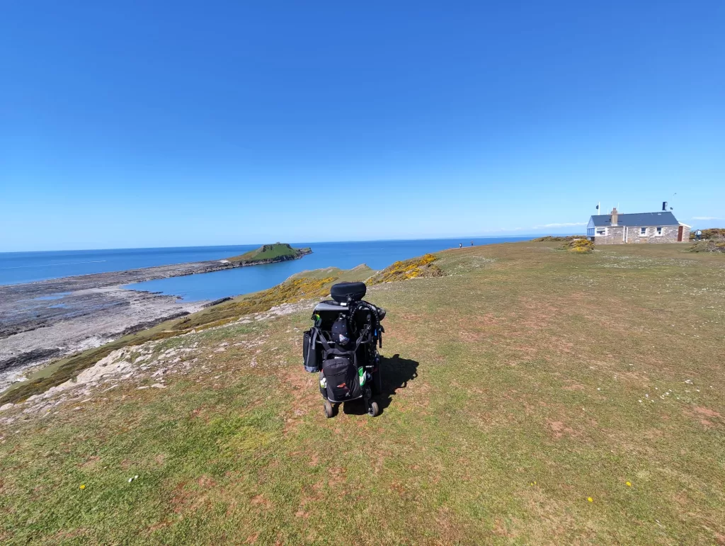 A wheelchair at Worms Head, with a view of the sea and coastal landscape, including a small island and a distant house under a clear blue sky.