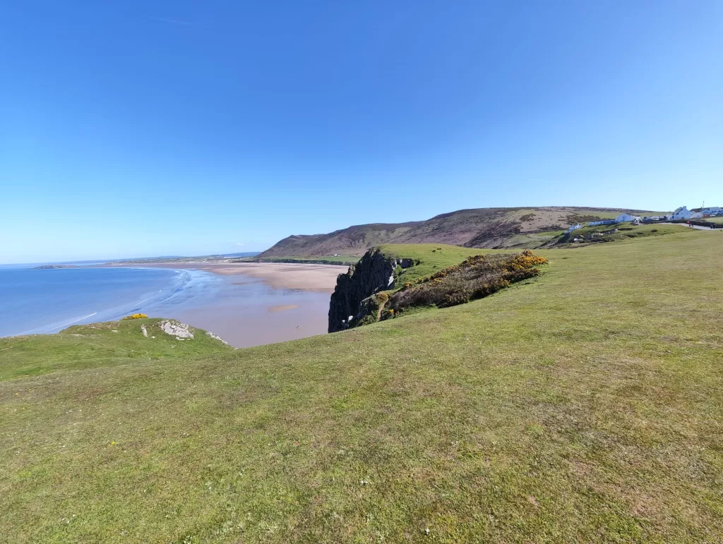 Panoramic view of Worms Head and Rhossili Bay, showcasing coastal scenery with a clear blue sky and lush green grass in the foreground.