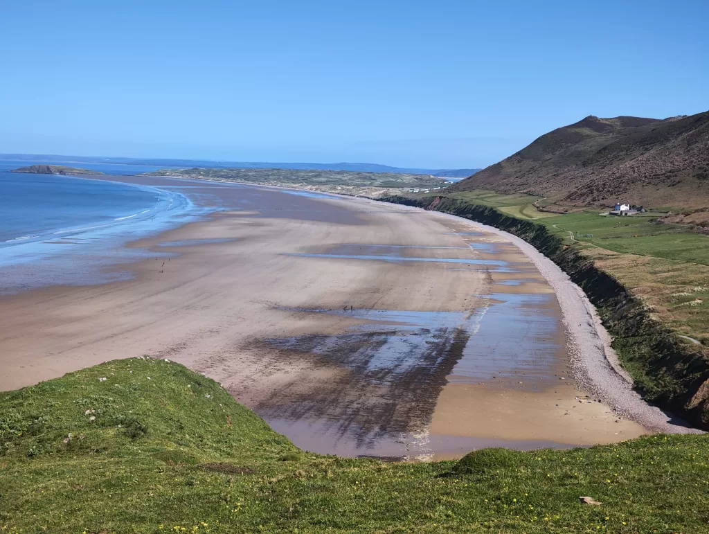 A panoramic view of Rhossili Bay on the Gower Peninsula, featuring a sandy beach and green cliffs under a clear blue sky.