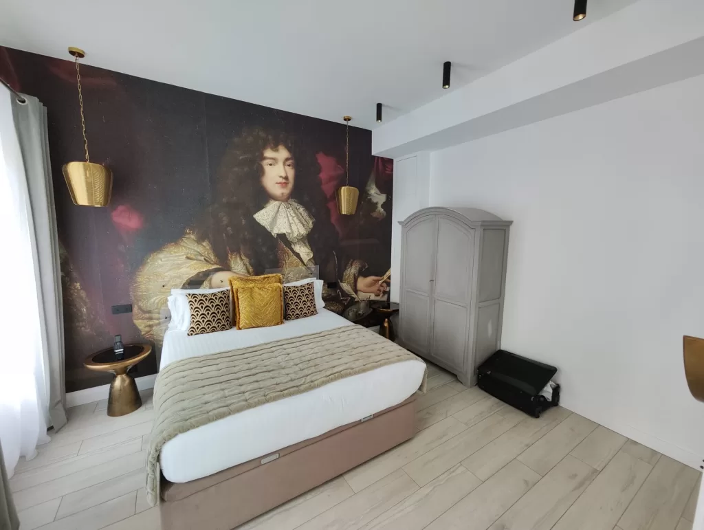 A stylish hotel room featuring a large bed adorned with decorative pillows, a mural of a historical figure on the wall, and modern furnishings including a small table and wardrobe.