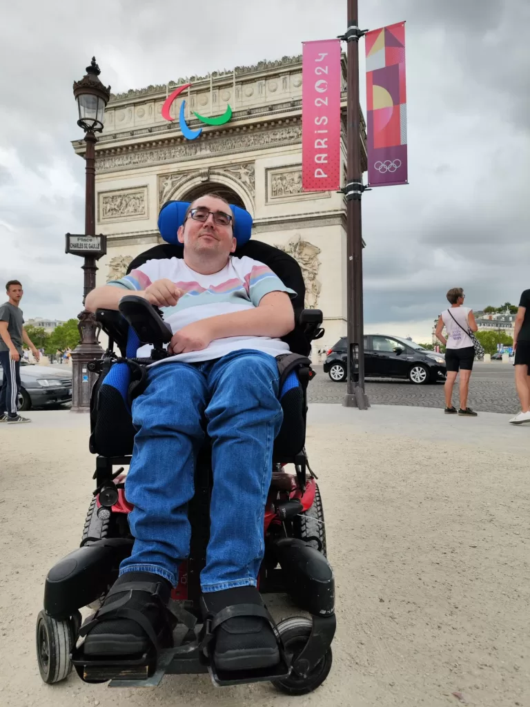 A person in a wheelchair smiling in front of the Arc de Triomphe in Paris, with banners for the 2024 Olympic Games in the background.