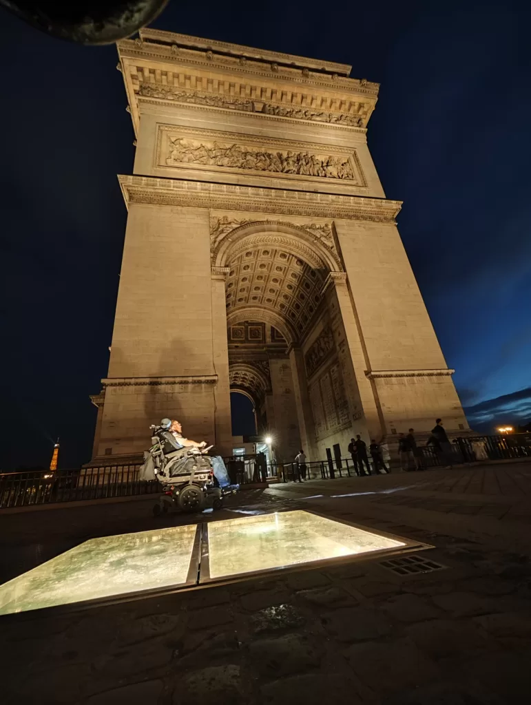 A person in a wheelchair is positioned in front of the illuminated Arc de Triomphe at night, with ambient city lights visible in the background.