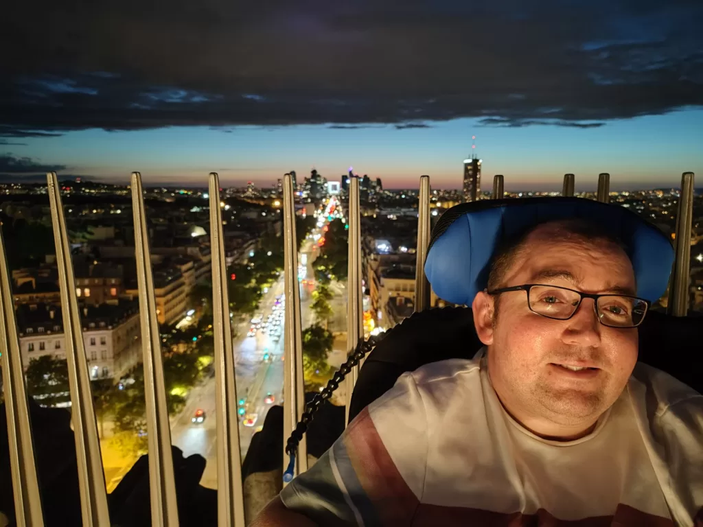 A person seated on a wheelchair enjoying the view of a city at night from a high vantage point, with a sunset in the background.