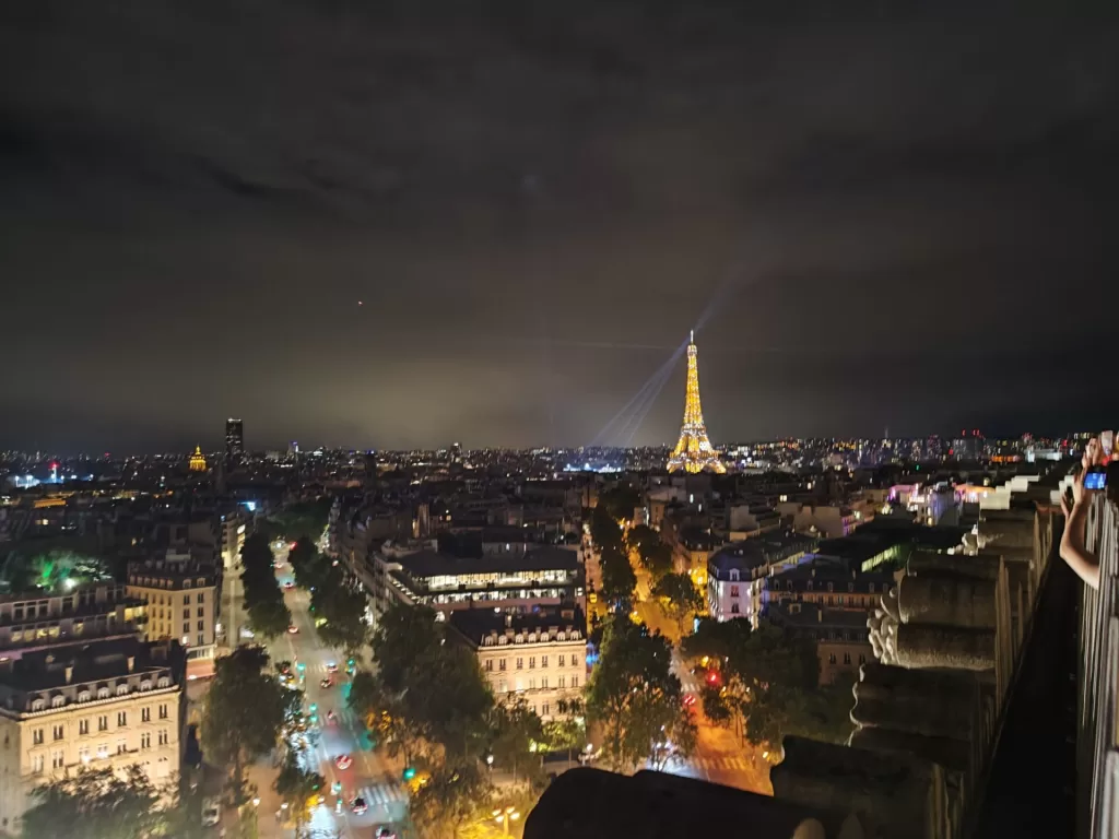 A nighttime view of Paris with the Eiffel Tower illuminated and beams of light shining from its top, showcasing the city's skyline and busy streets below.