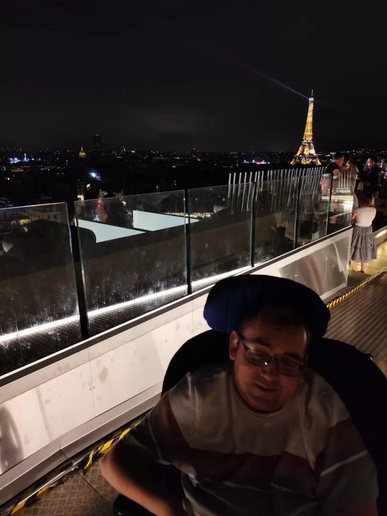 A person in a wheelchair with a blue headrest is seated on top of the Arc de Triomphe, overlooking a nighttime cityscape with the Eiffel Tower illuminated in the background.