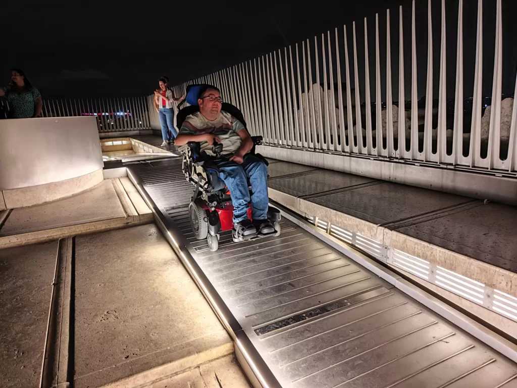 A person in a wheelchair enjoying the view from a raised platform at night on top of the Arc de Triomphe, surrounded by a railing and with other visitors in the background.