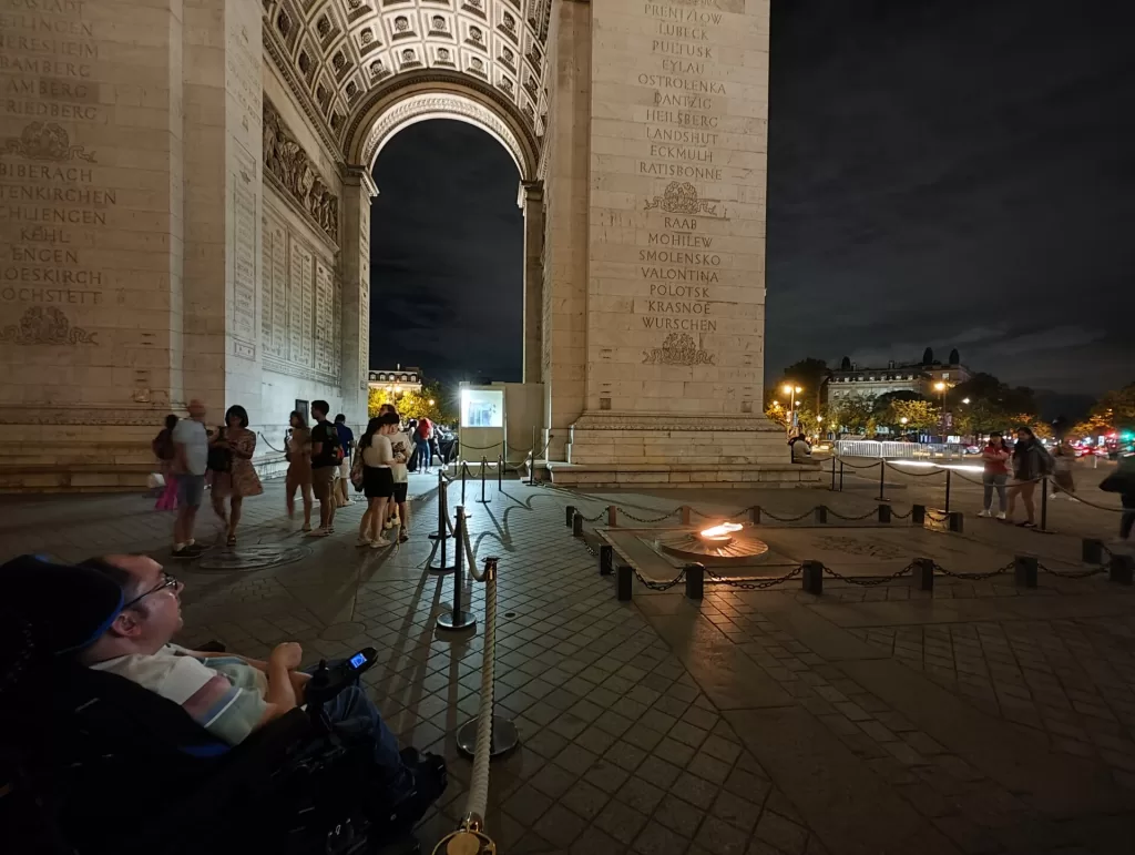 A person in a wheelchair observes the Arc de Triomphe at night, with people walking nearby and a flame at the base of the monument.