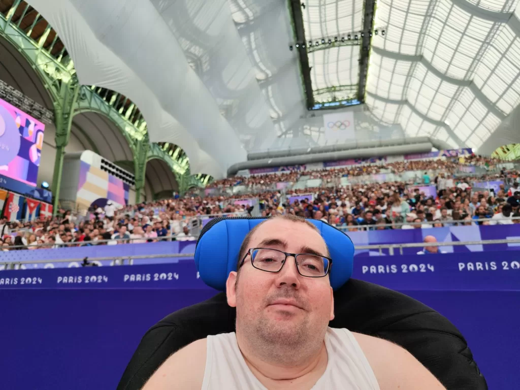 A person in a wheelchair with a serene expression is seated in the Grand Palais during the Olympic Games in Paris, surrounded by a large audience under a grand, glass-domed roof. The vibrant Paris 2024 branding is visible in the background.