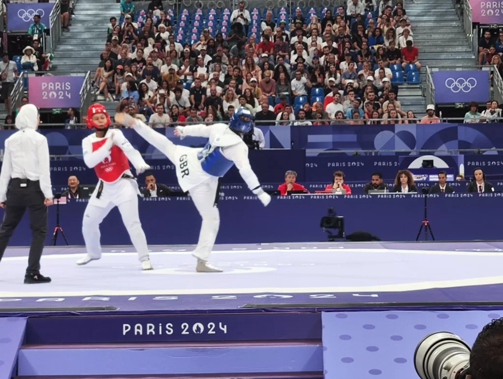Two taekwondo competitors engage in a match at the Paris 2024 Olympics, with one athlete in a red uniform and the other in blue. Spectators are visible in the background.