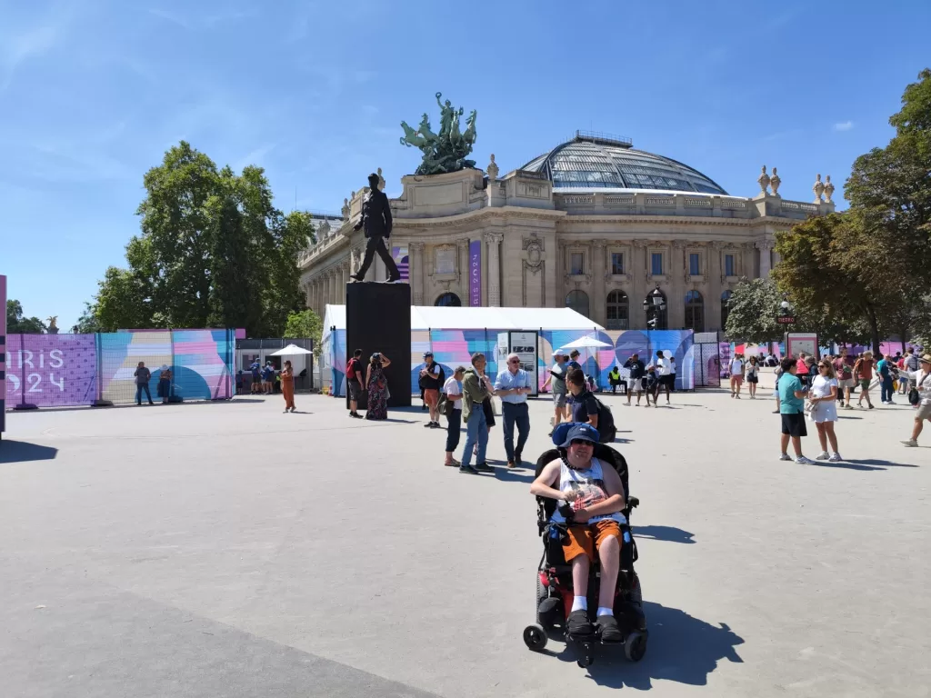 A person in a powered wheelchair is sitting in the foreground of a bustling outdoor area, with people walking around and a large venue building in the background, featuring a statue and colourful signage for the Paris 2024 Olympics.