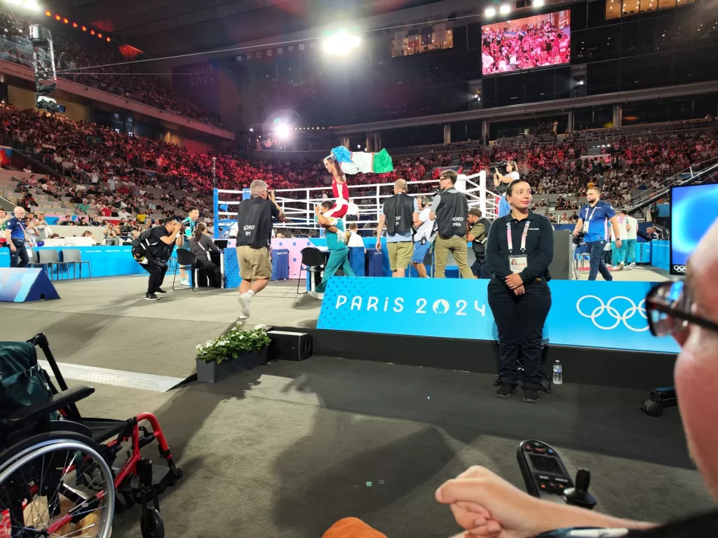 View of the boxing ring at the 2024 Paris Olympics with spectators in the background, wheelchair visible in the foreground.