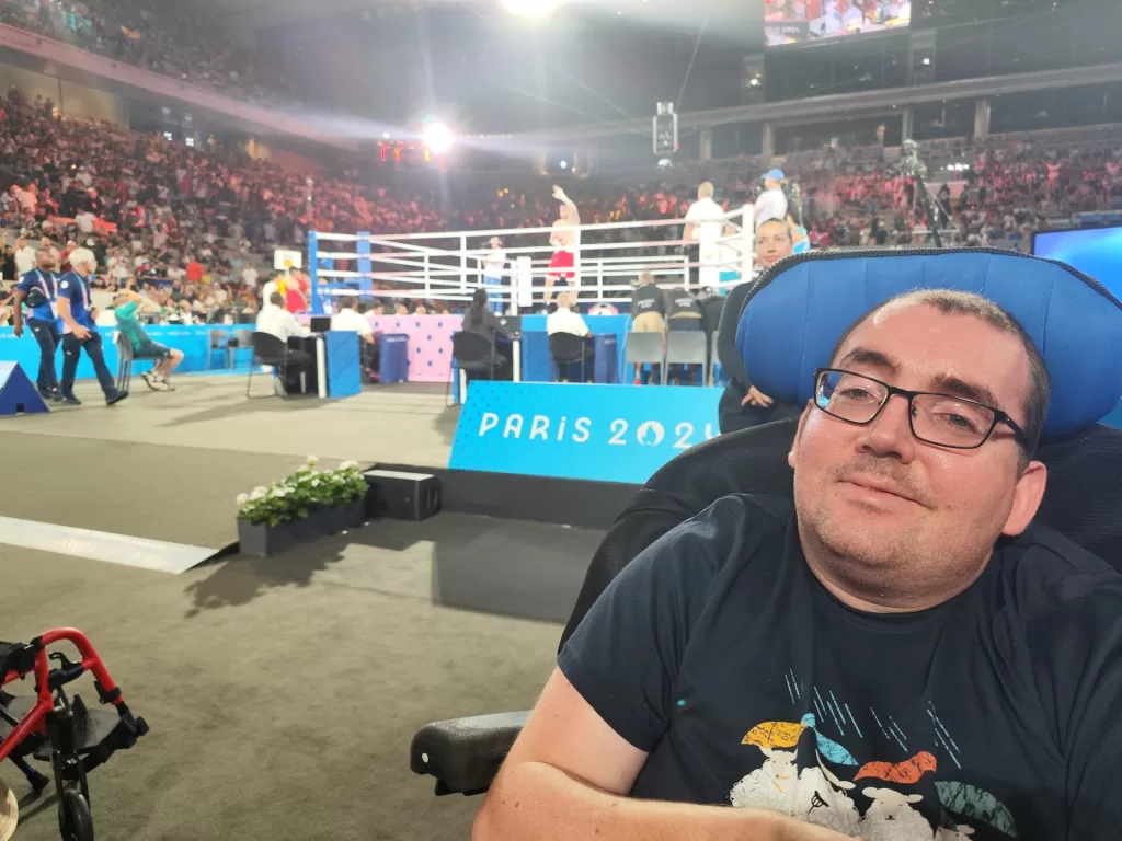 A spectator in a wheelchair at the boxing event during the Paris 2024 Olympics, with the boxing ring and an audience in the background.
