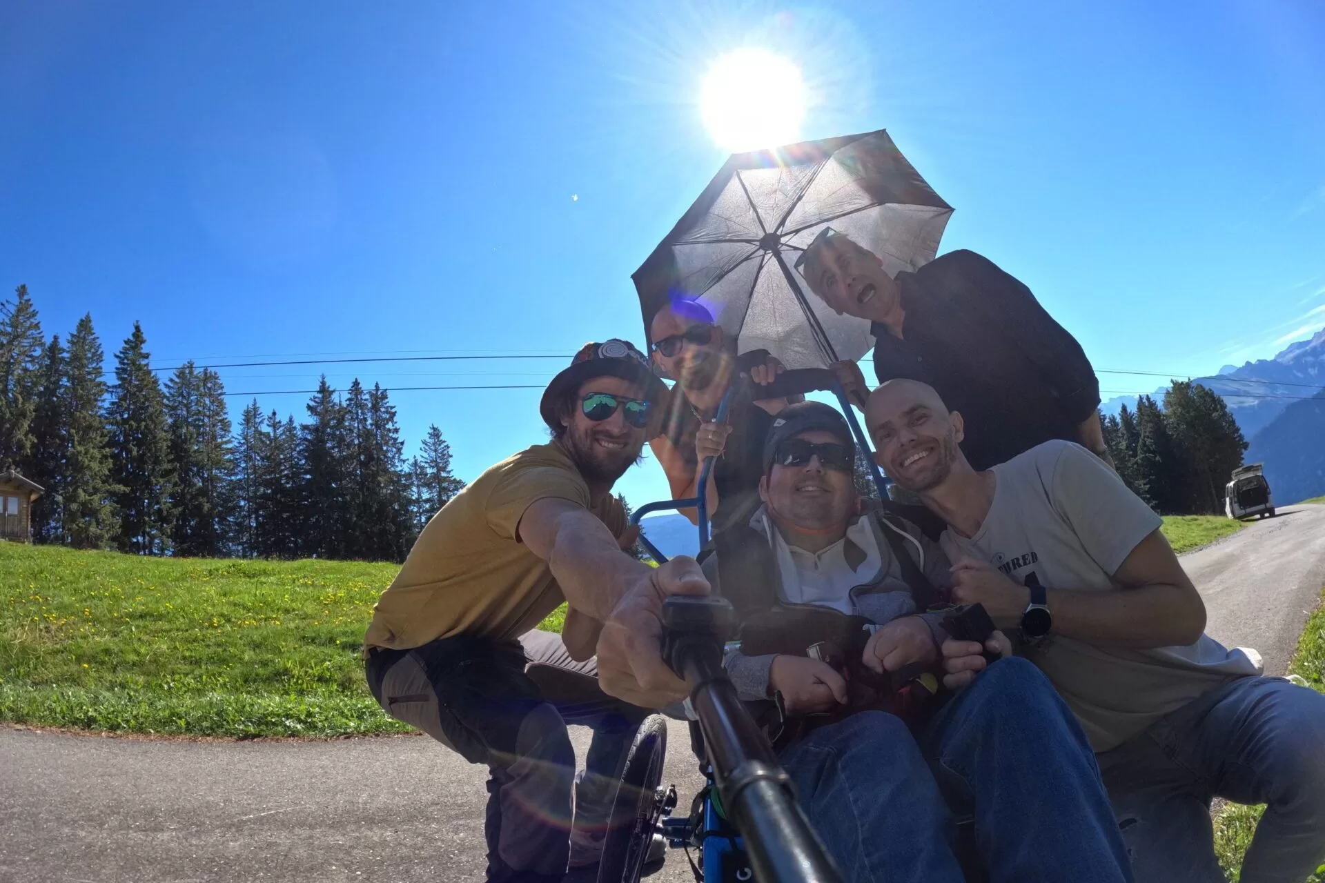 A group of five people smiling and posing for a selfie outdoors, with one person in a wheelchair holding an umbrella, set against a bright blue sky and green trees.