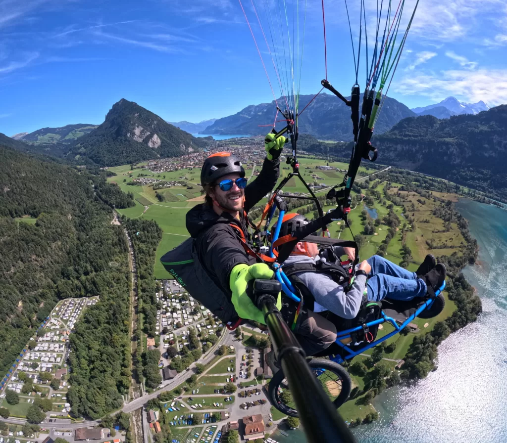 A thrilling paragliding experience over a scenic landscape in Switzerland, featuring a person smiling while suspended in the air, with green fields, mountains, and a lake visible below.