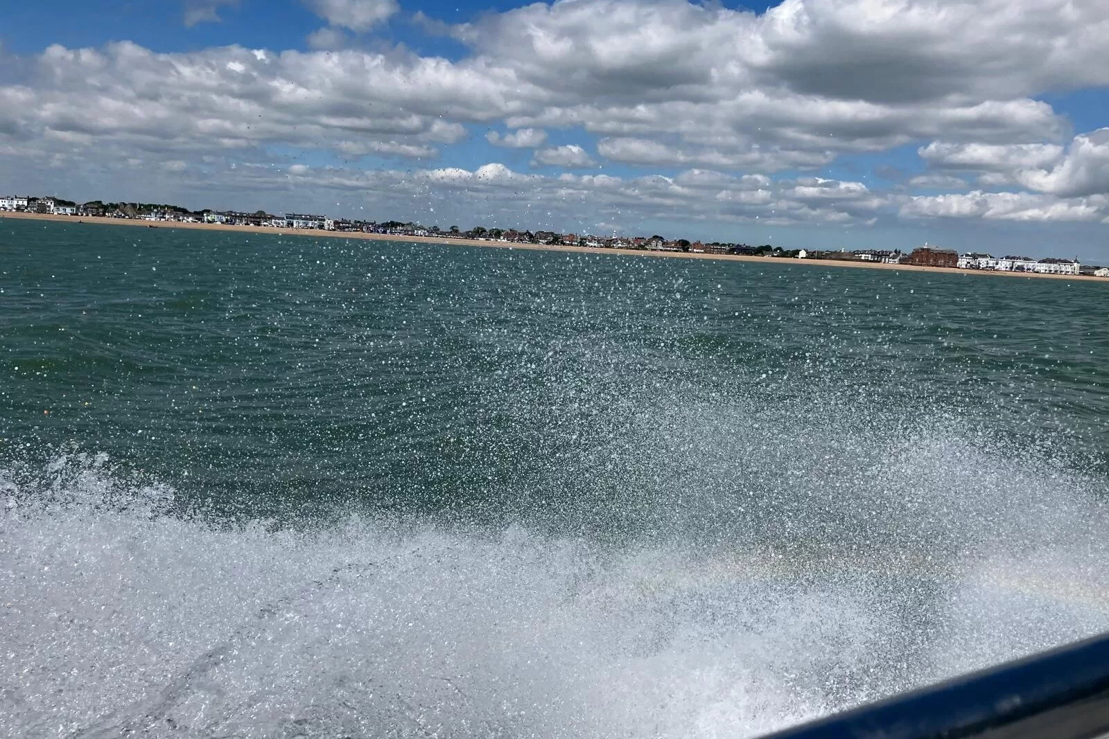 A view from a boat, showing water splashing as the boat moves quickly, with a beach and buildings in the background under a partly cloudy sky.