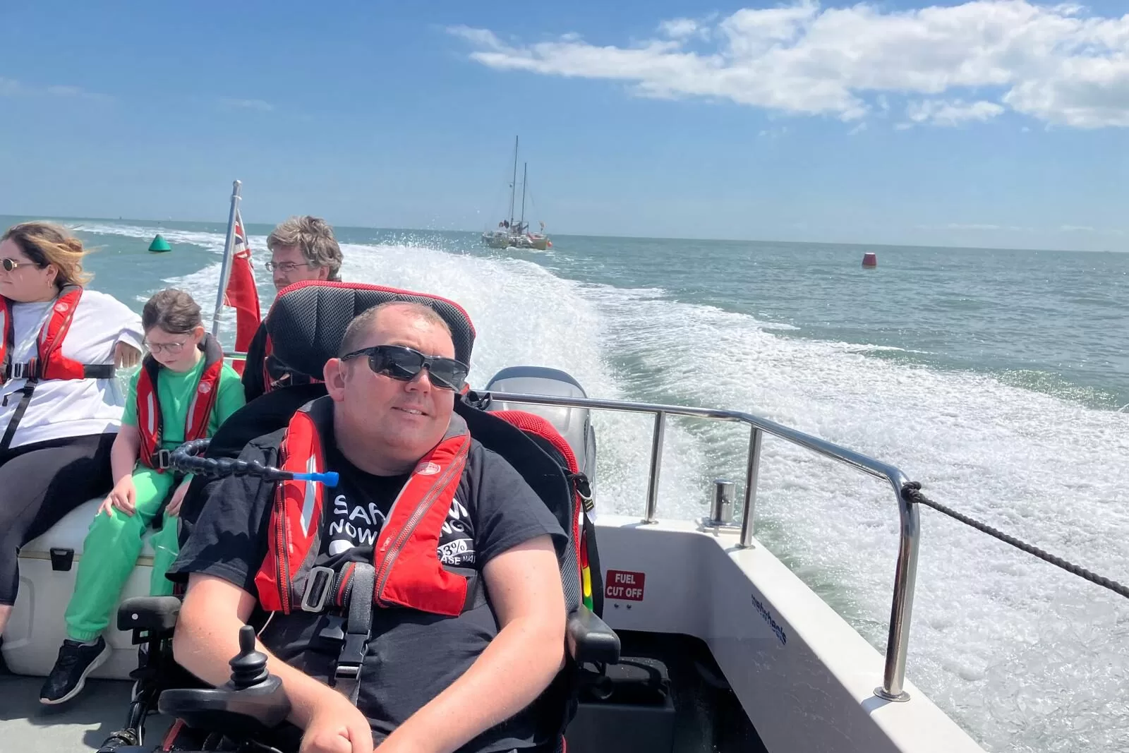 A group of people enjoying a boat trip on a sunny day, with one individual in a wheelchair equipped with a life jacket.