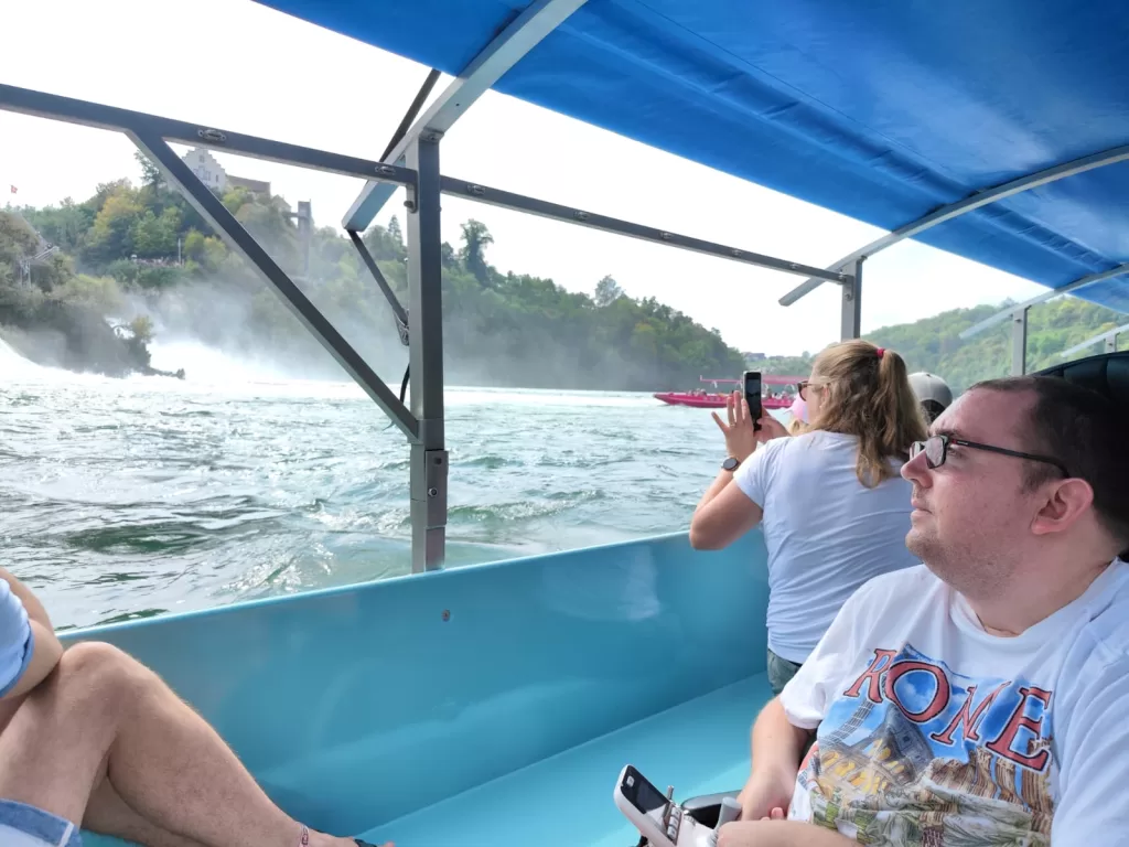 Passengers on a boat approaching the Rhine Falls, with one person taking a photo while another enjoys the view.