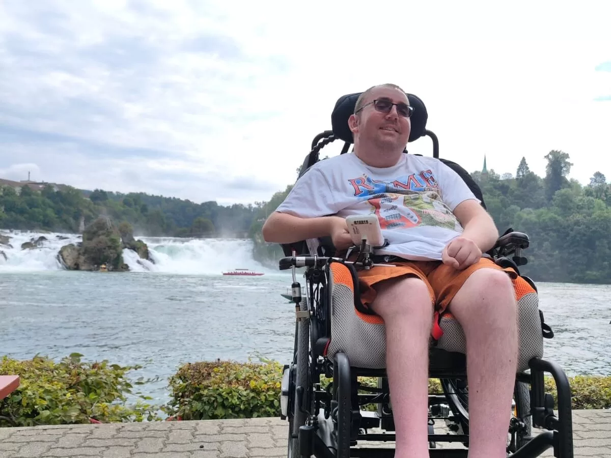 A person in a wheelchair smiles while seated by the Rhine Falls in Switzerland, with the waterfall and trees visible in the background.