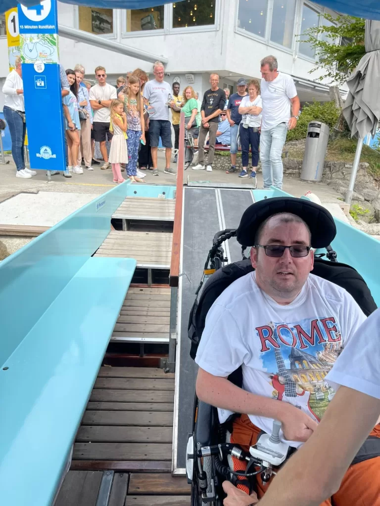 A man in a wheelchair is preparing to board a boat, with a group of people observing nearby. The boat has a ramp for accessibility.