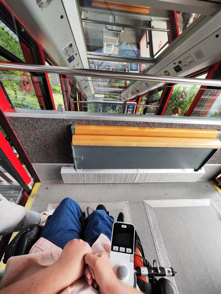 View from inside a funicular railway, showing a passenger in a wheelchair. The railway features red walls and a scenic view through the transparent windows.