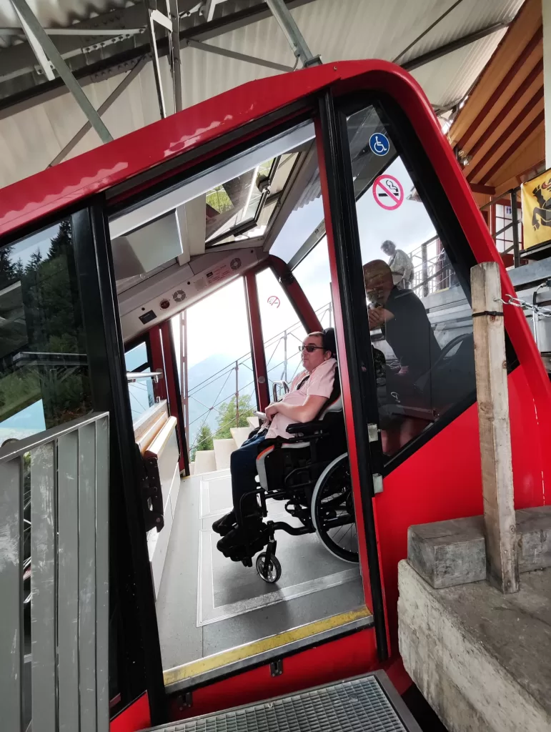 A person in a wheelchair inside a red cable car, with an accessible entrance and views of the surrounding area.