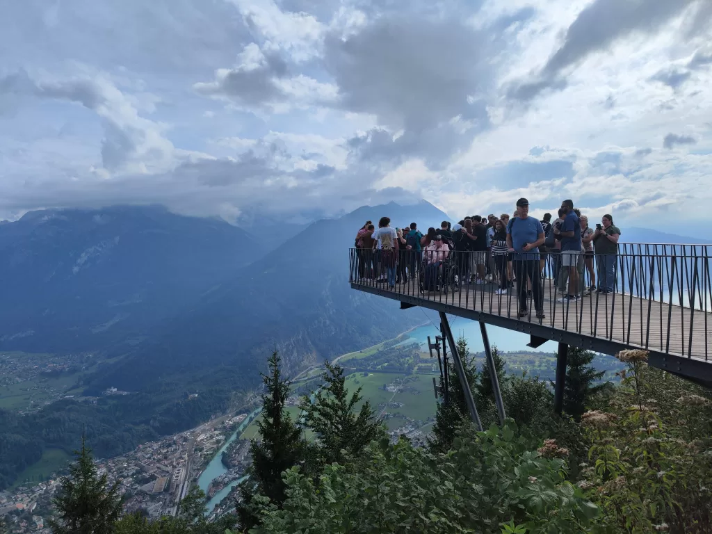 A crowded viewing platform on a mountain overlooking a valley with mountains in the background and a cloudy sky.