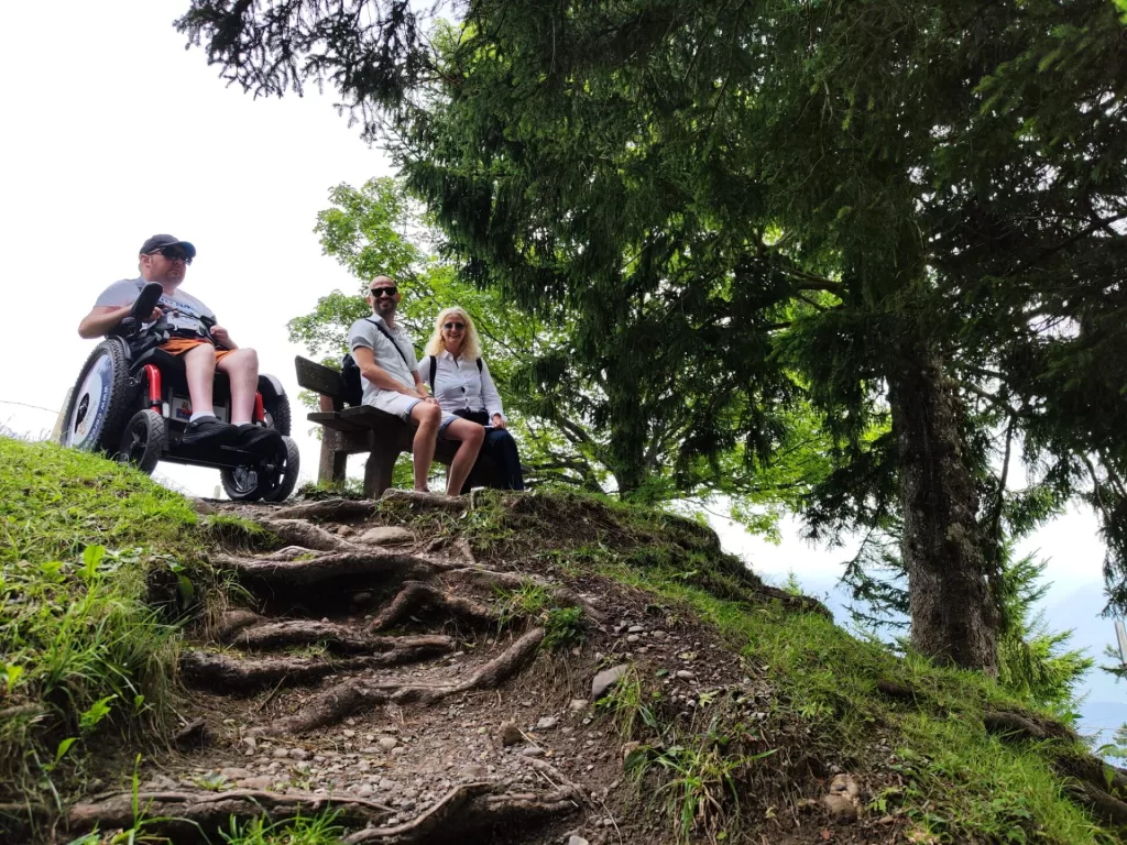 A person in a power wheelchair sitting on a grassy slope, with two individuals seated on a bench nearby, surrounded by trees.