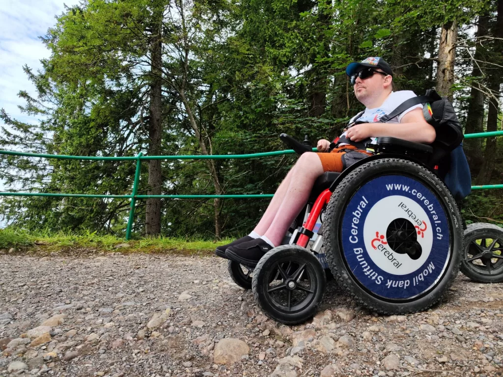 A person in a wheelchair equipped with large wheels, sitting on a gravel path surrounded by trees, enjoying a scenic view.