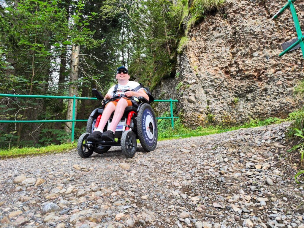 A person sitting in an all-terrain wheelchair on a gravel path in a forested area, with trees and rocky terrain in the background.
