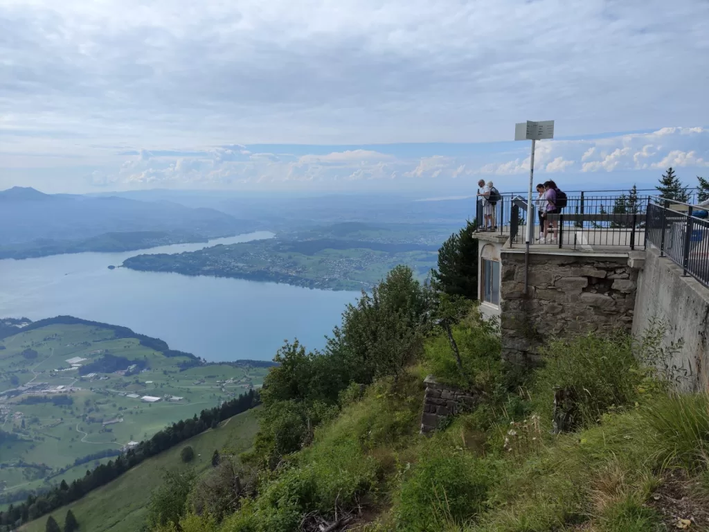 Panoramic view from a mountain overlook showing a lake surrounded by green hills and distant mountains under a cloudy sky.