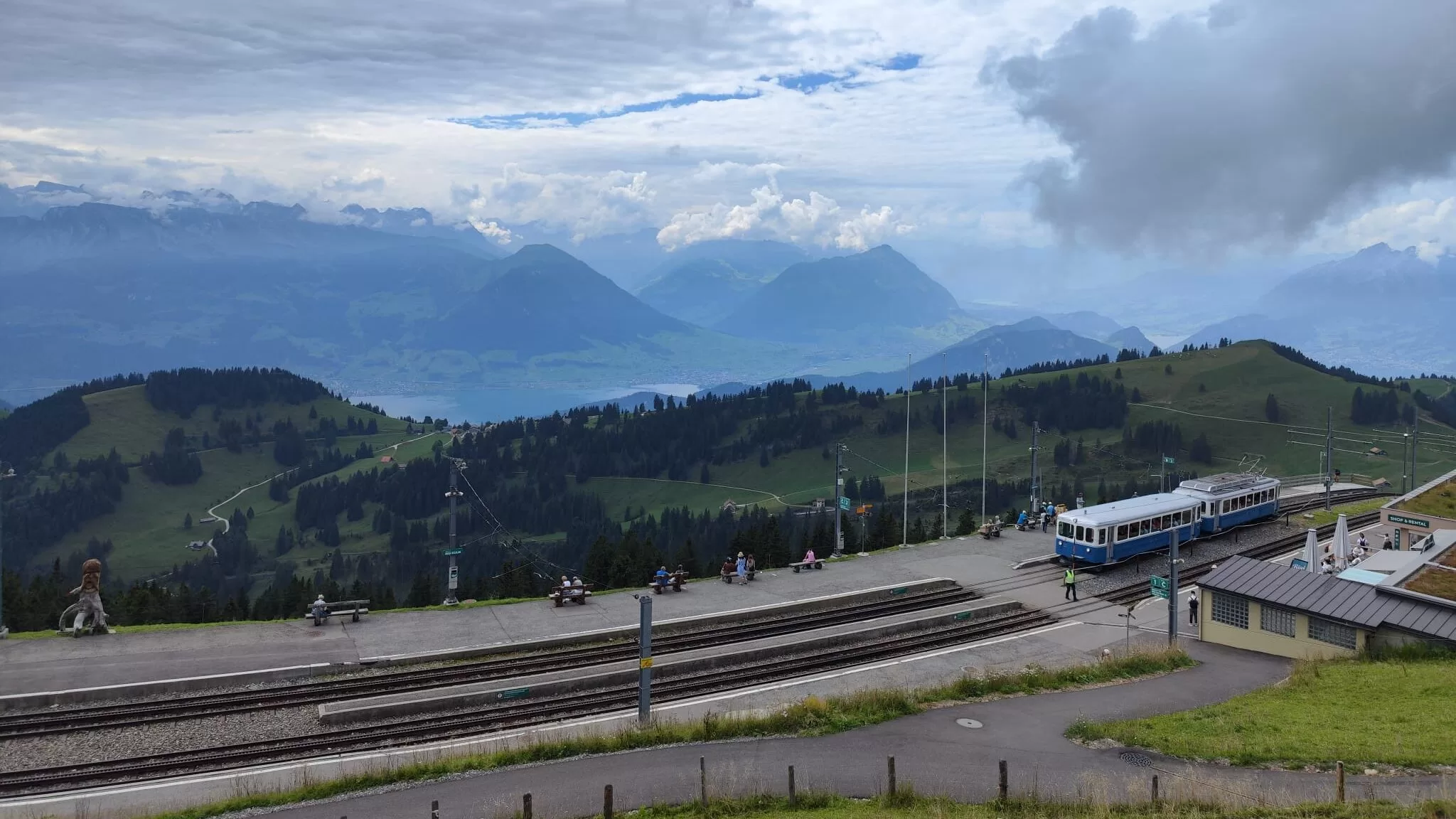 A panoramic view of the Swiss mountains and valleys from a train station, with green hills, a railway track, and a group of people enjoying the scenery.