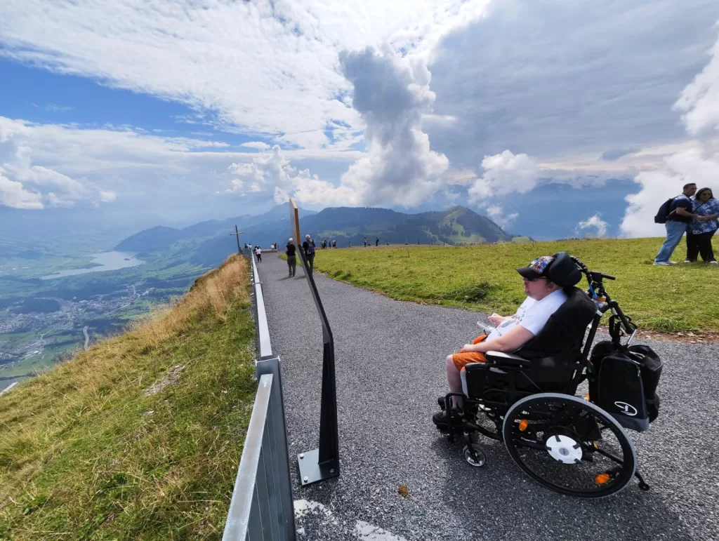A person in a wheelchair enjoying a scenic view from a mountain summit, with lush green hills and cloudy skies in the background.