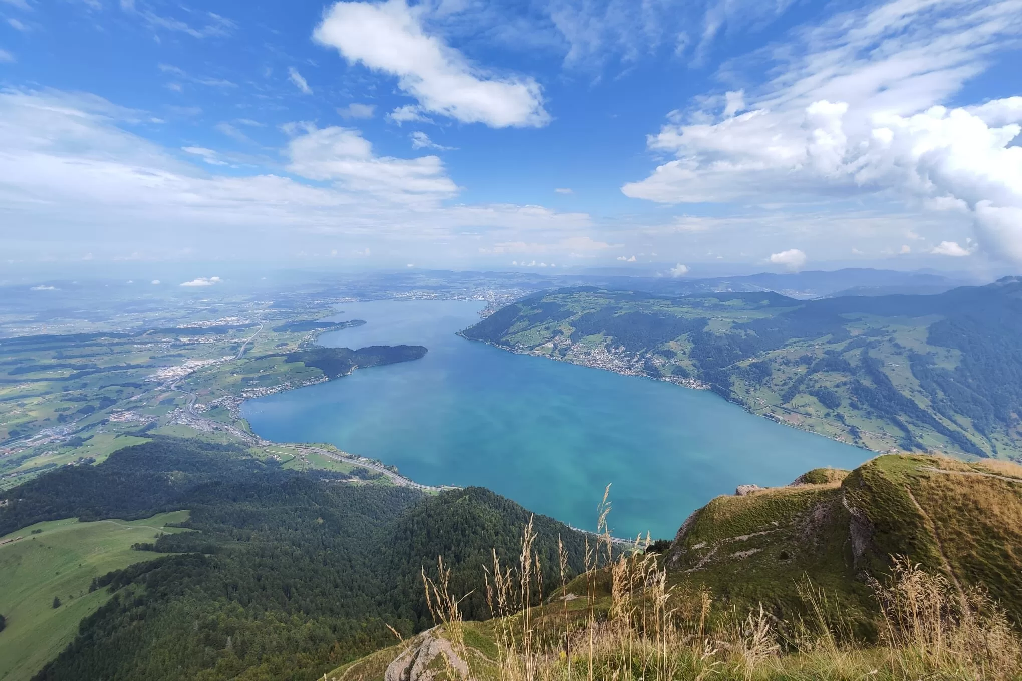 A panoramic view of Lake Lucerne from a mountain, featuring lush green hills and blue waters under a partly cloudy sky.