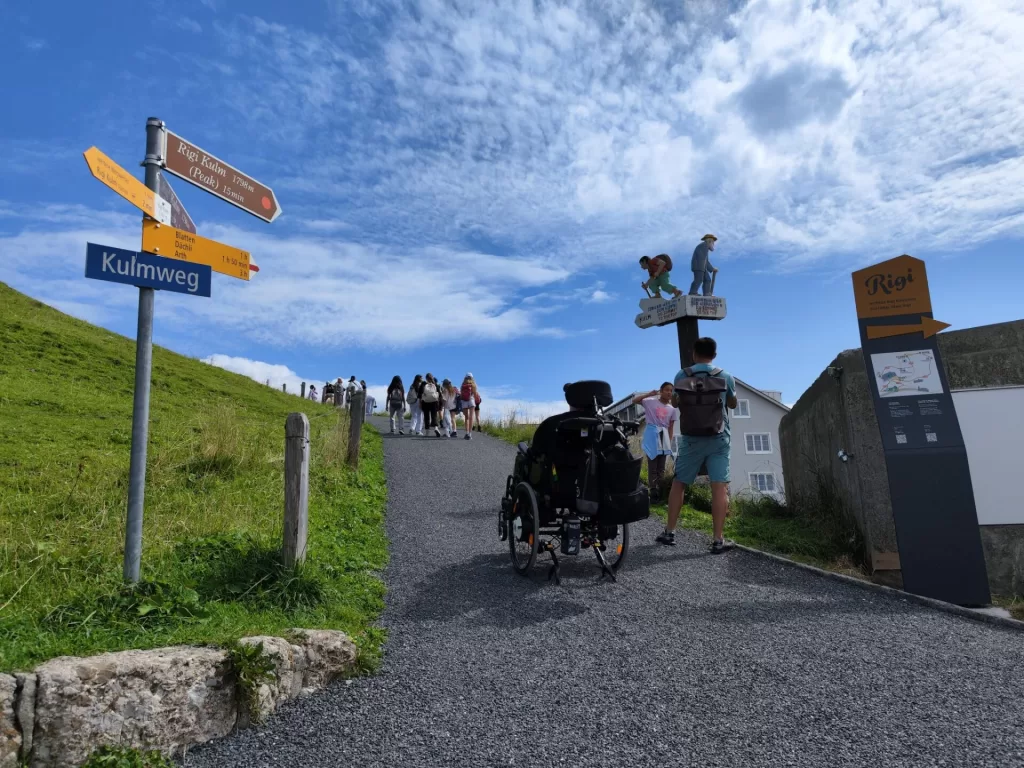 A pathway leading up to the summit of Mount Rigi, with directional signs indicating the peak and accessible routes. A person in a wheelchair is sitting on the path, accompanied by others walking towards the viewpoint.
