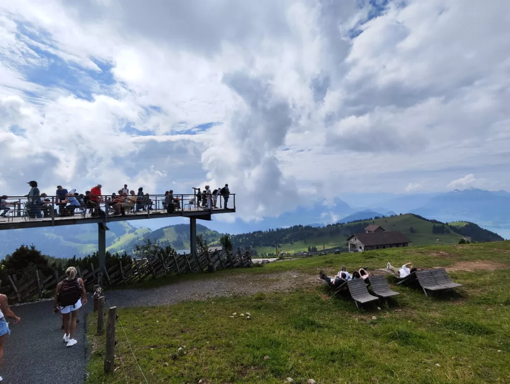 A scenic view from a mountain lookout point, showing people enjoying the outdoors on a wooden deck with picturesque hills and clouds in the background.
