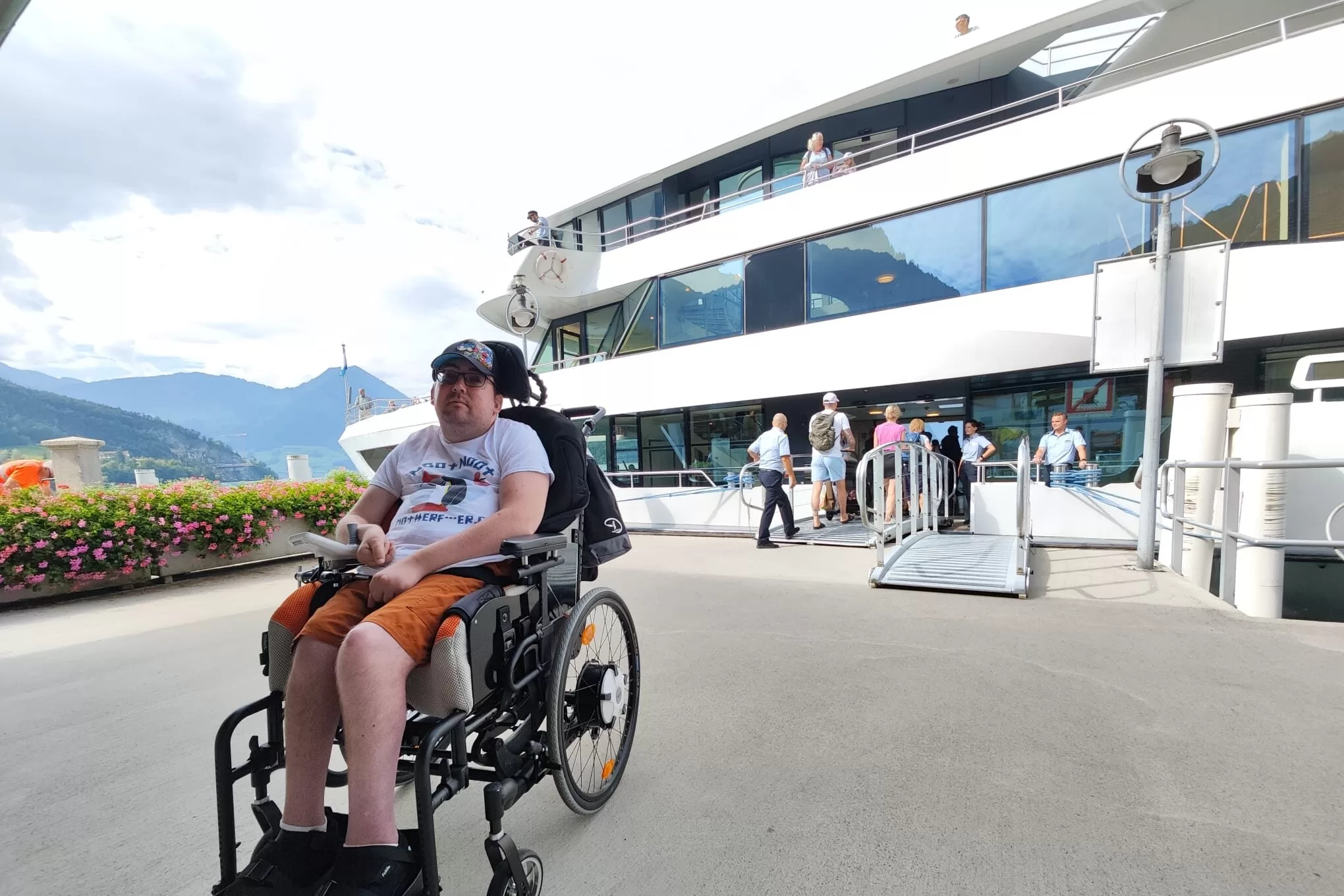 A person in a wheelchair sits on the dock next to a boat. Behind them, a group of people is boarding the boat. The scenery includes mountains and flowers along the dock.