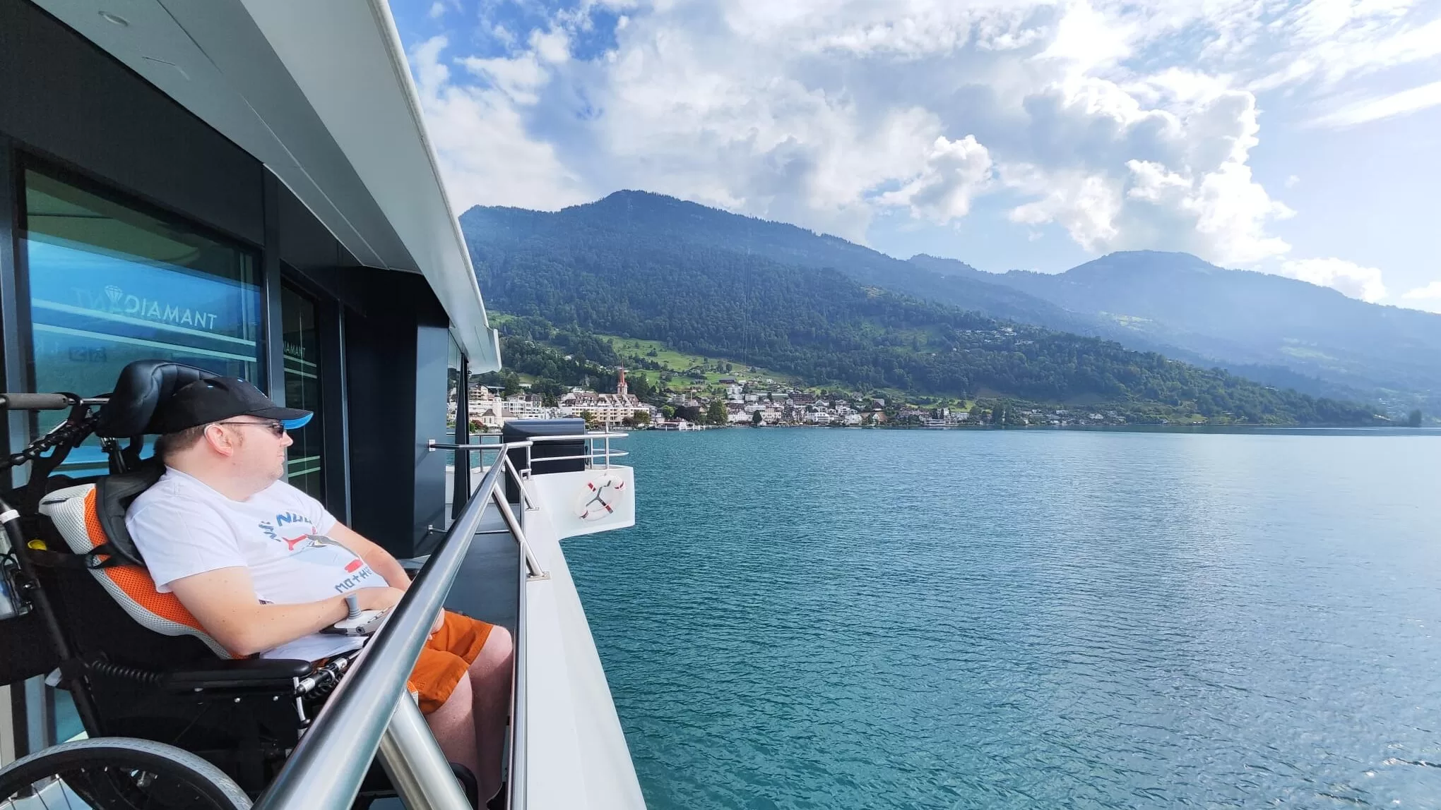 A person in a wheelchair enjoying the view from a boat on a lake, surrounded by mountains and a picturesque village.