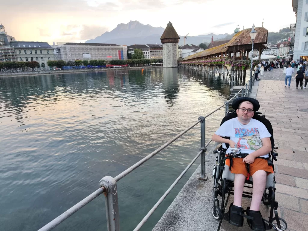 A person in a wheelchair sits beside Lake Lucerne with the iconic Chapel Bridge and mountains in the background.