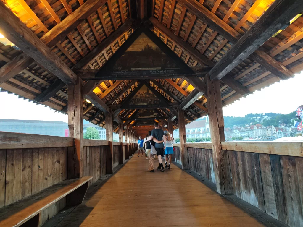 A view of the interior of Chapel Bridge in Lucerne, showcasing its wooden structure and walkway with people walking through.