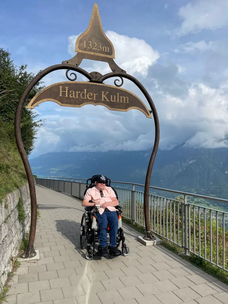 Person in a wheelchair at the Harder Kulm viewpoint sign, which indicates an elevation of 1323 meters, with mountain scenery in the background.