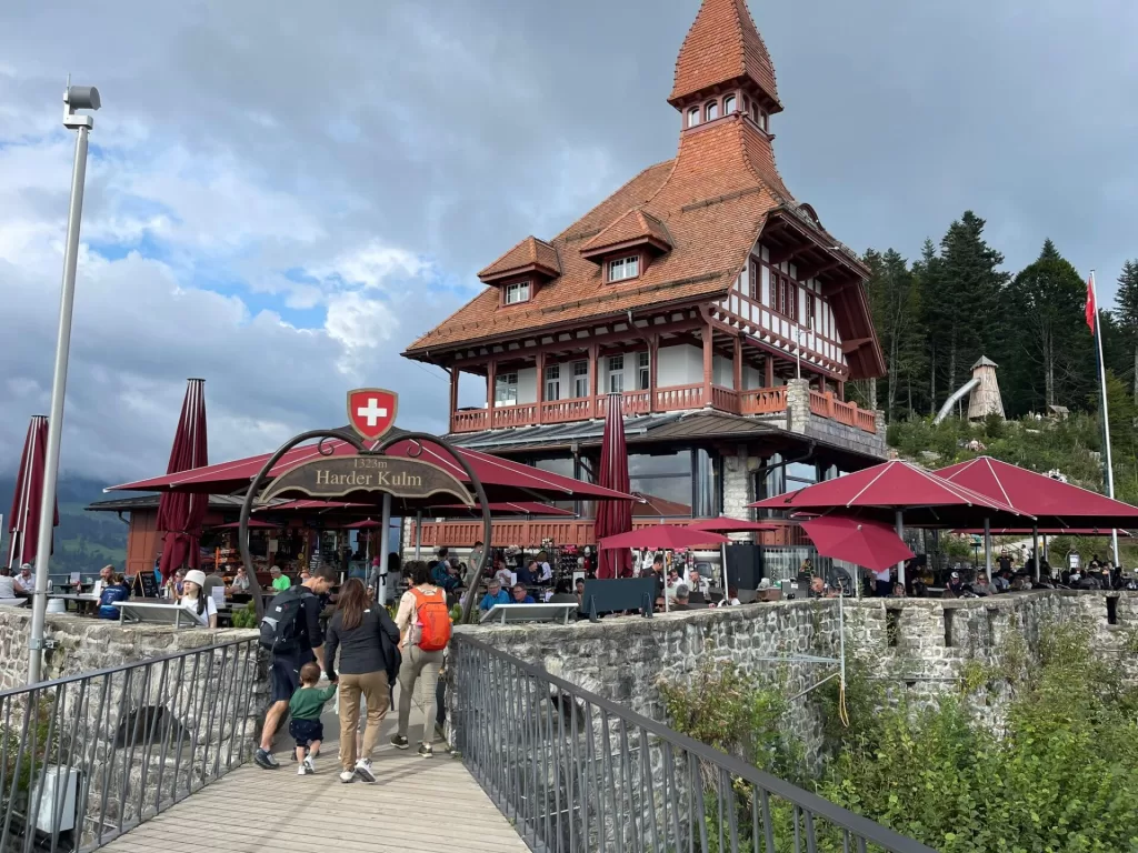A scenic view a mountain restaurant at the top of Harder Kulm in Switzerland, featuring a distinctive wooden building with a steep red roof, surrounded by guests enjoying their time on the terrace and the scenic landscape.